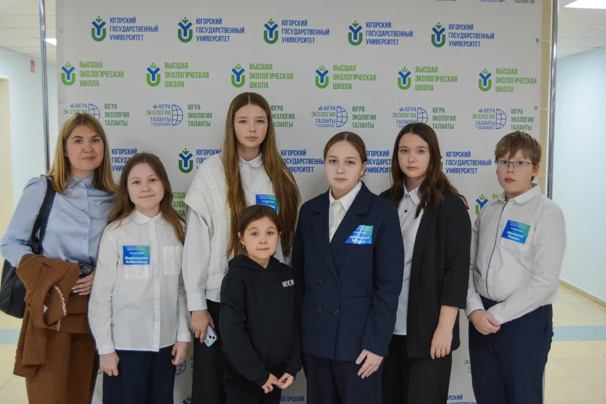 На фотографии группа из восьми человек стоит на фоне баннера. На баннере повторяется логотип 