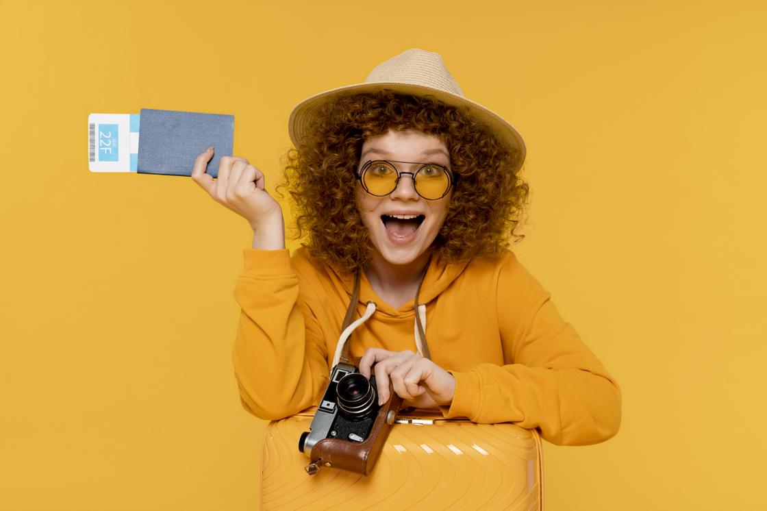 Кучерявая девушка с чемоданом и фотоаппаратом держит в руках билеты