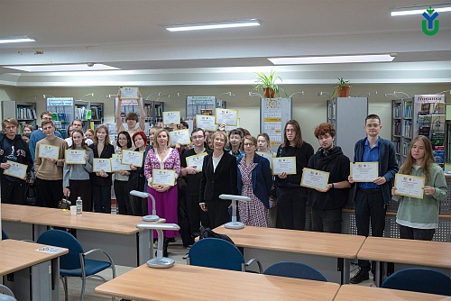На изображении показана группа людей, стоящих в библиотеке. Люди держат сертификаты участника. На заднем плане расположены книжные полки, заполненные книгами. На полках видны таблички с надписями 