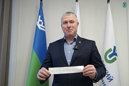 На фотографии изображен мужчина средних лет, стоящий в помещении. Он одет в синий пиджак и рубашку в мелкую клетку. К пиджаку прикреплен беспроводной микрофон Godox. На фоне видны три флага: синий с белым символом, белый и белый с зеленым и синим символом