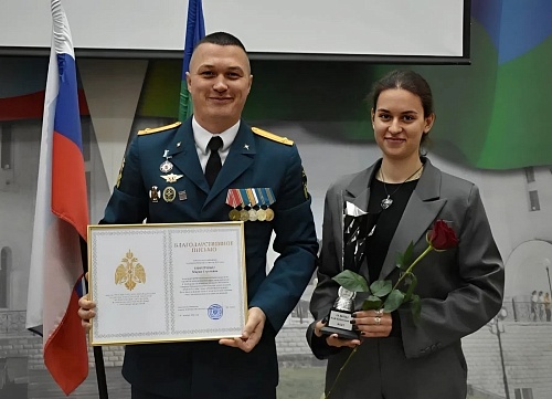 На фотографии изображены два человека: мужчина в военной форме и девушка в сером пиджаке. Мужчина стоит слева, он держит в руках рамку с текстом, в которой можно различить слова 