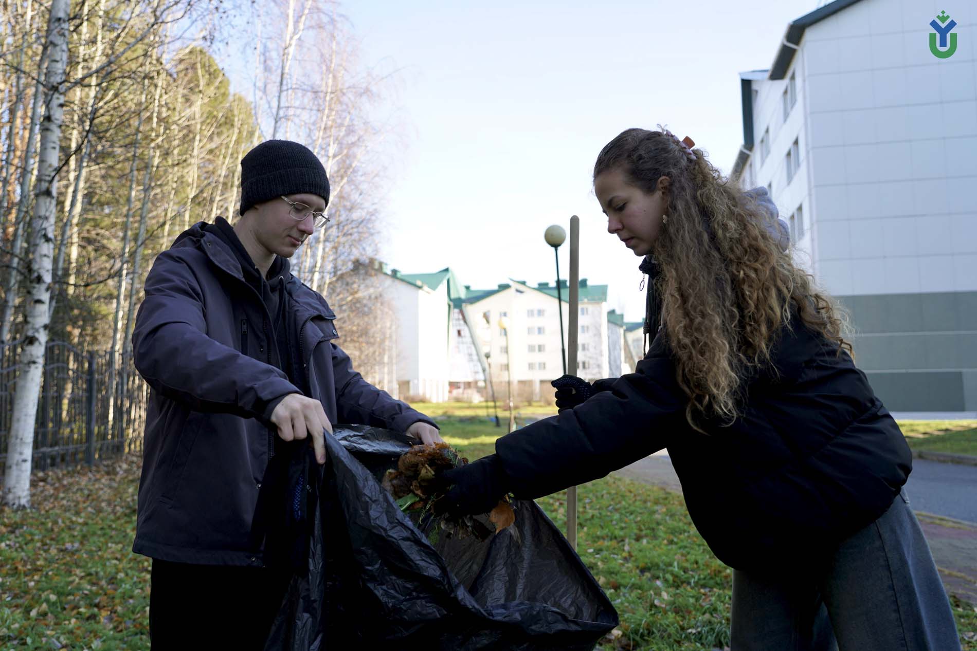 На изображении два человека собирают листья в черный мусорный мешок. Молодой человек слева одет в черную вязаную шапку, очки, темно-синюю куртку и черную толстовку. Он держит верхнюю часть мусорного мешка. Девушка справа имеет длинные вьющиеся волосы, соб