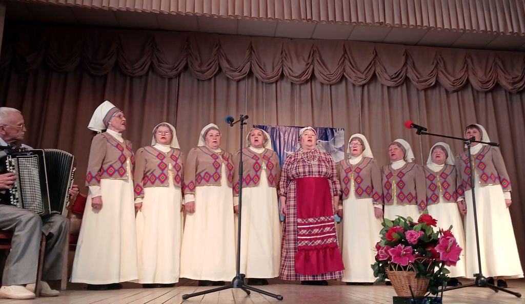 На сцене хор из девяти взрослых женщин в национальных костюмах, слева мужчина - аккомпаниатор.