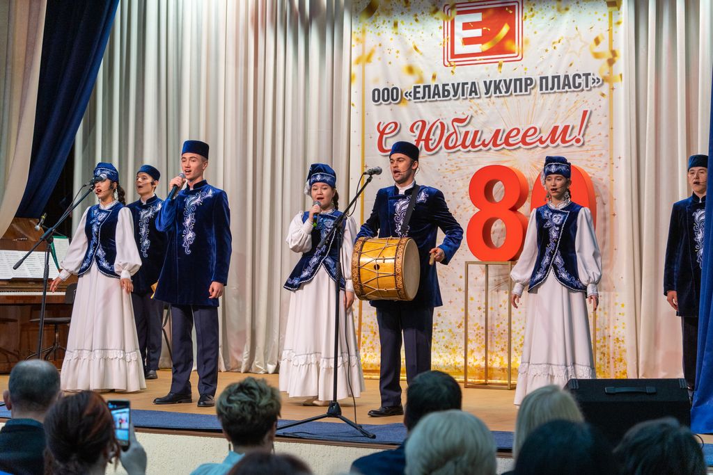 На сцене коллектив художественной самодеятельности - молодые люди в татарских национальных костюмах бело-синего цвета. На заднем плане баннер с надписью: 