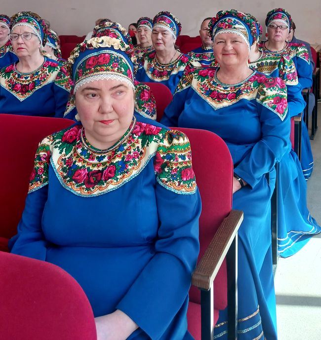 В зрительном зале сидят участницы женского хора в синих национальных сарафанах и расшитых головных уборах.